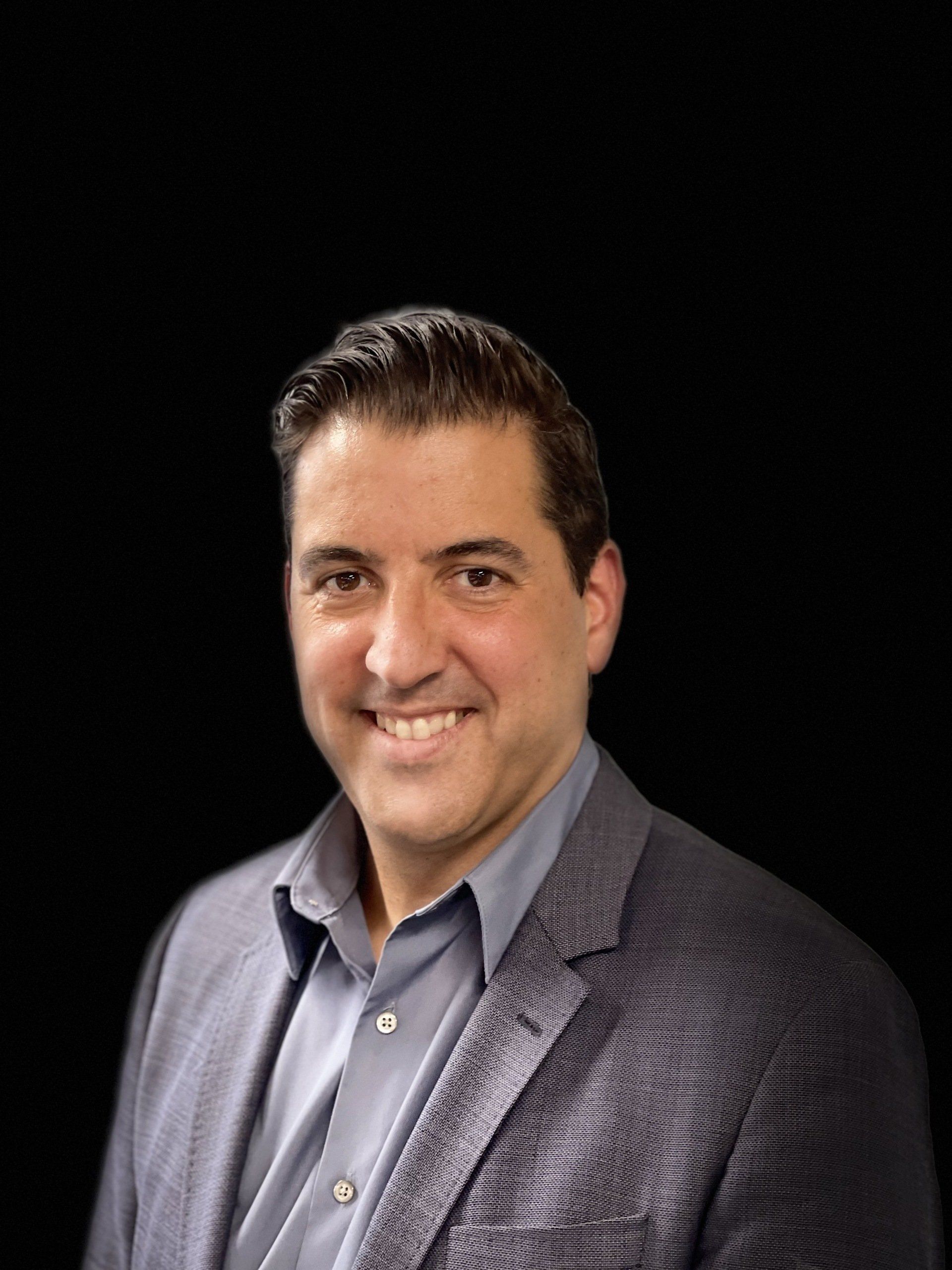A man in a suit and blue shirt is smiling in front of a black background.