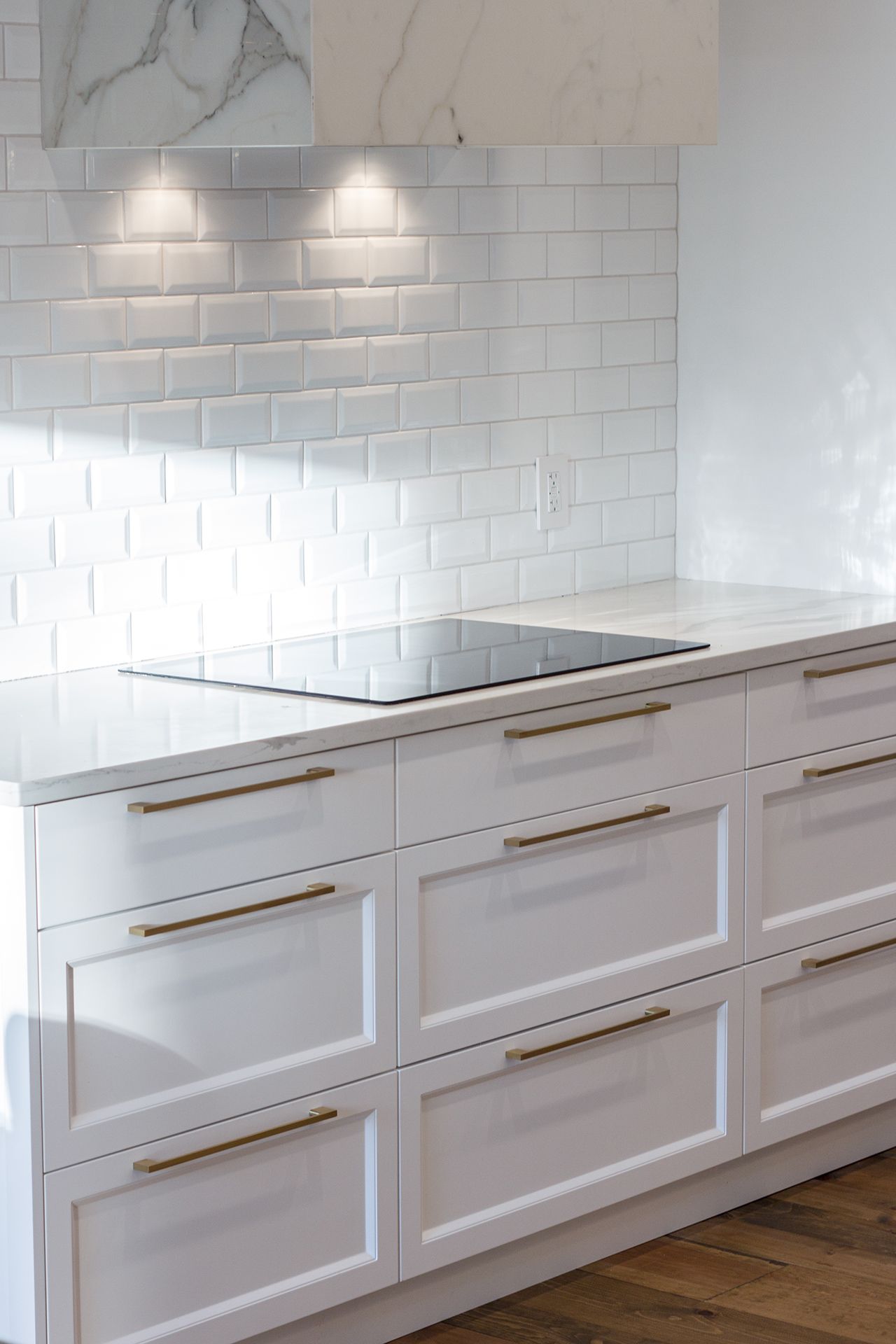 Cuisine blanche avec cuisinière, placards blancs à poignées dorées et crédence en carrelage blanc.