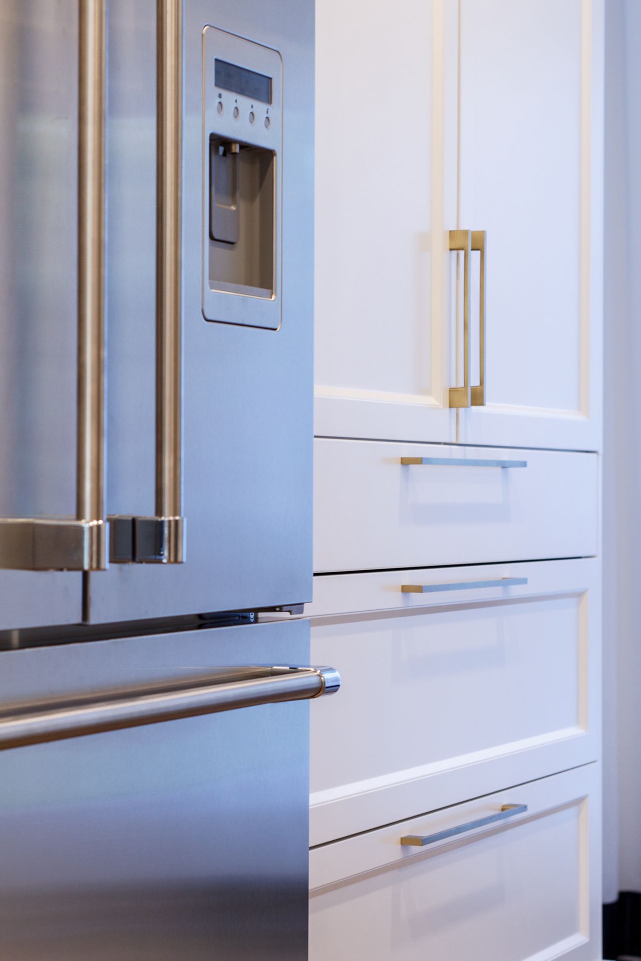 Réfrigérateur en acier inoxydable à côté d'armoires de cuisine blanches avec quincaillerie dorée.