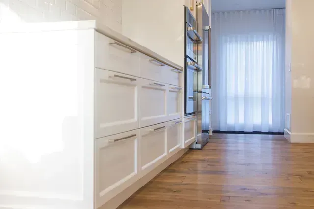 Armoires de cuisine blanches avec poignées argentées, plancher de bois franc et une fenêtre avec des rideaux transparents.