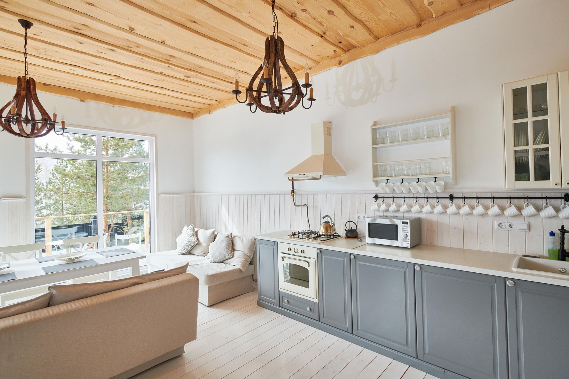 Kitchen with grey cabinets, white walls, wooden ceiling, two chandeliers, and a window.