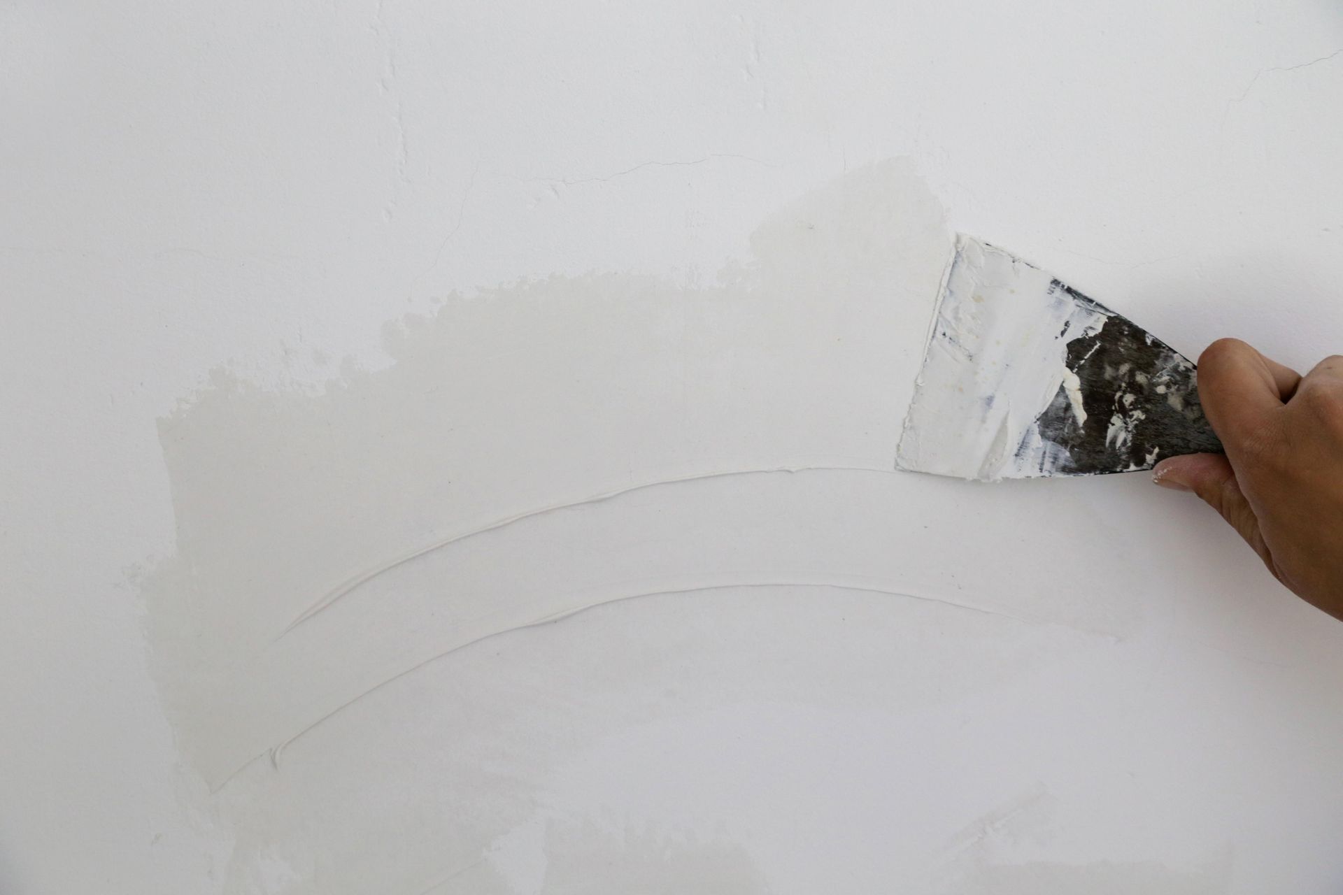 Hand using a putty knife to spread white joint compound on a white wall.