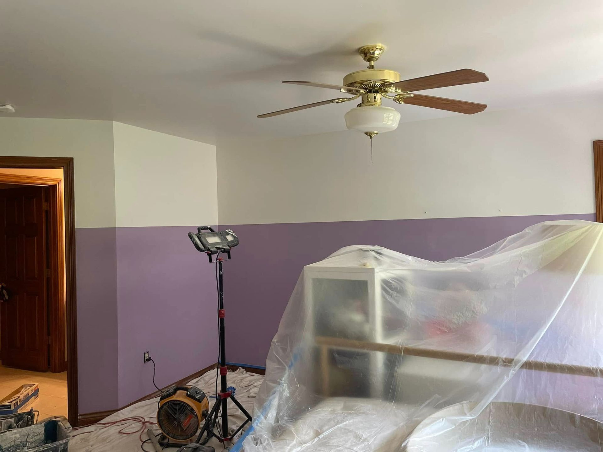 Room with half-painted lavender walls, covered furniture, ceiling fan, and work light.