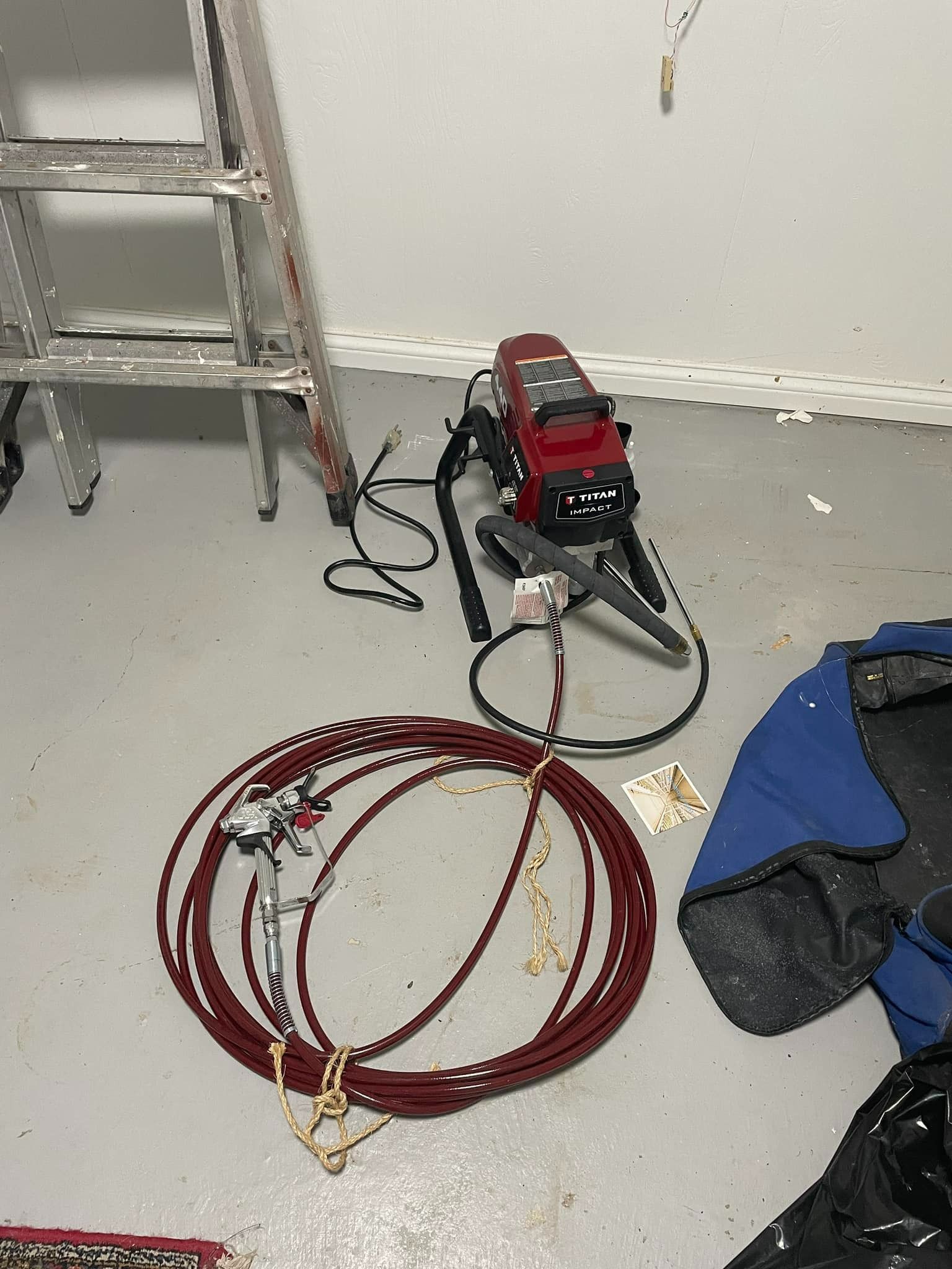 Ladder and paint sprayer on a floor. A red hose and gun are coiled nearby.