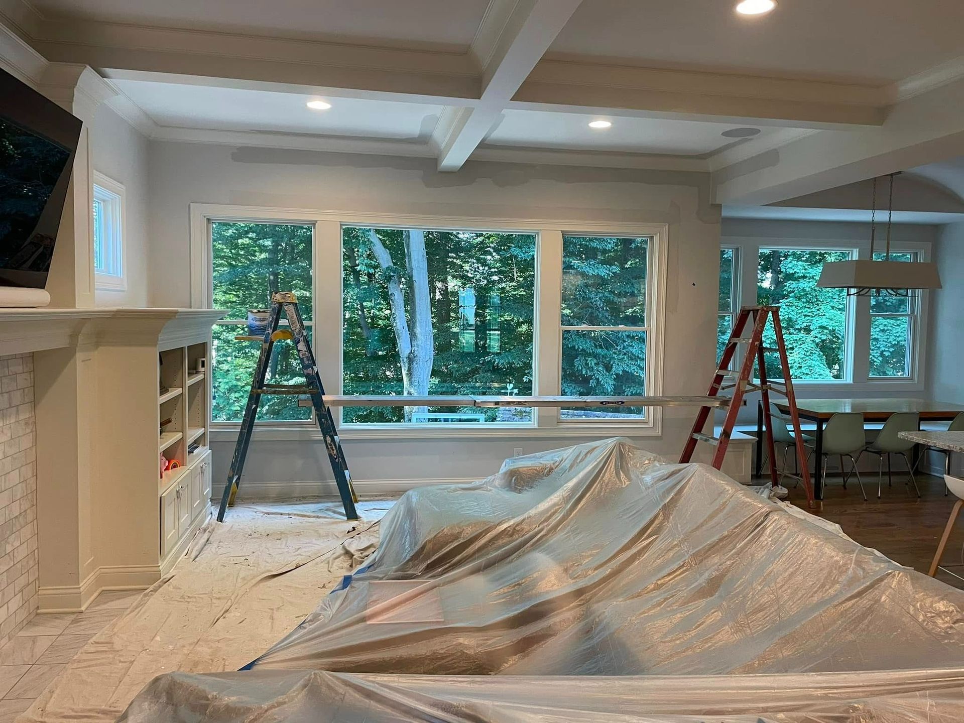 Room interior with paint supplies; furniture covered in plastic; ladders; windows; and white walls and beams.