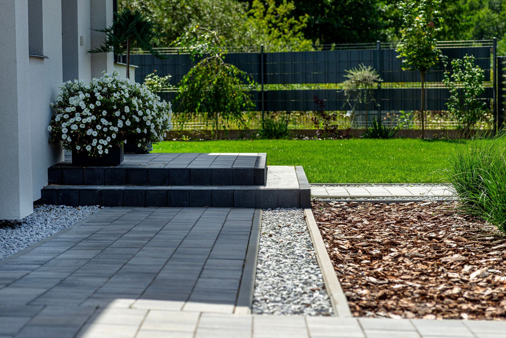 Gray stone patio with steps, flower pot, gravel, and bark, leading to a lawn with trees and a black fence.