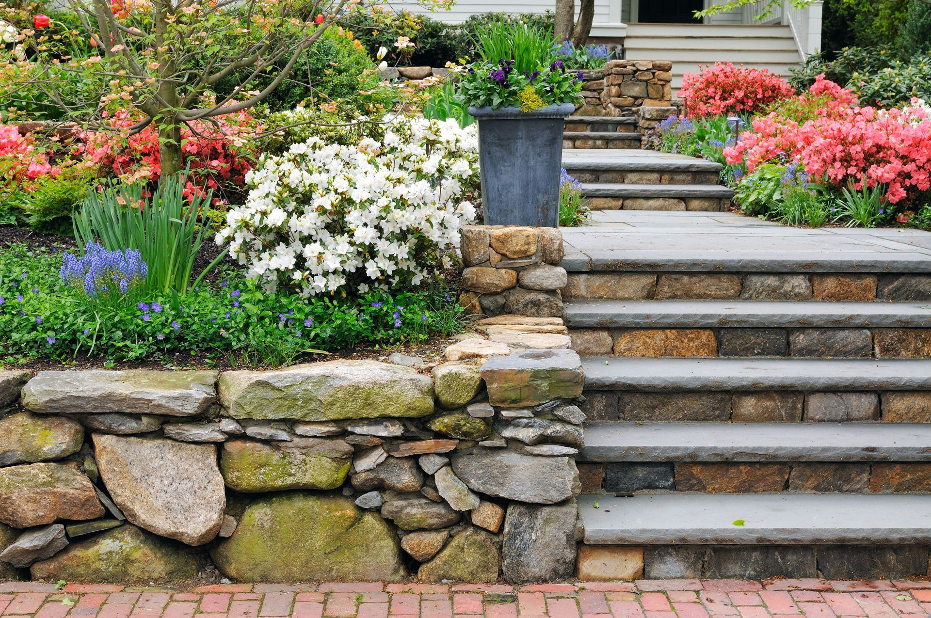 A stone wall with stairs leading up to a house surrounded by flowers