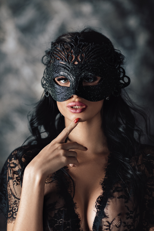 A person with dark hair wears an ornate black lace mask and matching lace top, posing with a finger near their lips.