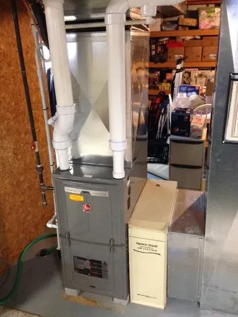 A grey high-efficiency furnace with vertical PVC venting pipes stands next to a tall, cream-colored air filter cabinet.