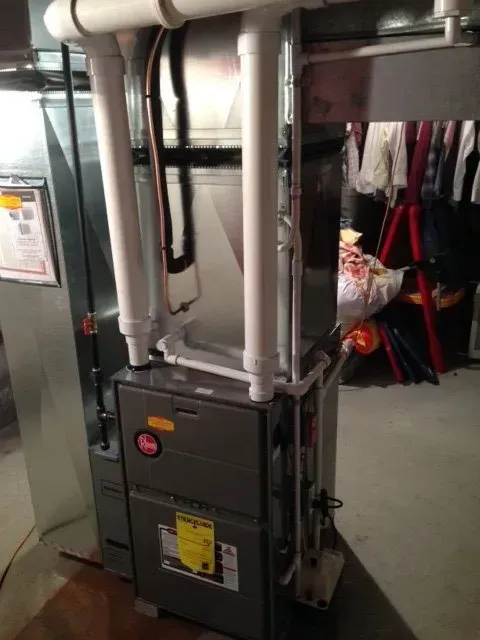 A gray high-efficiency furnace with vertical white PVC venting pipes in a basement utility area.