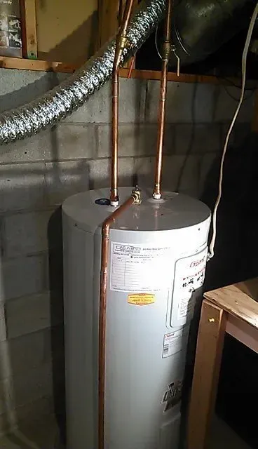 A white electric water heater stands against a concrete block wall with two vertical copper pipes extending from the top.
