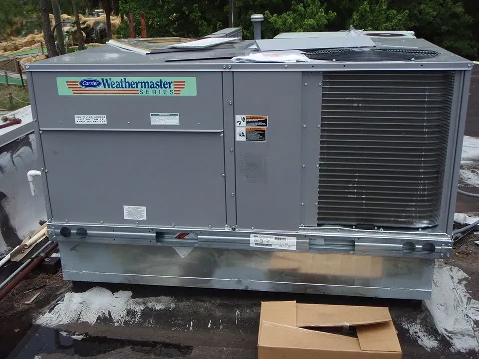 A gray Carrier WeatherMaster rooftop HVAC unit sitting on a flat, gravel-covered roof next to a cardboard box.