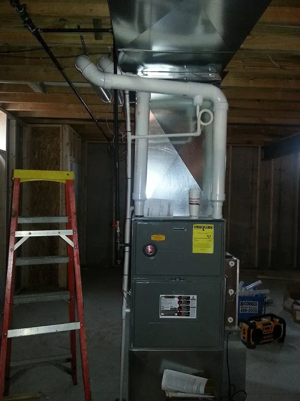 A grey furnace with white PVC exhaust pipes attached, standing in an unfinished basement beside a red stepladder.