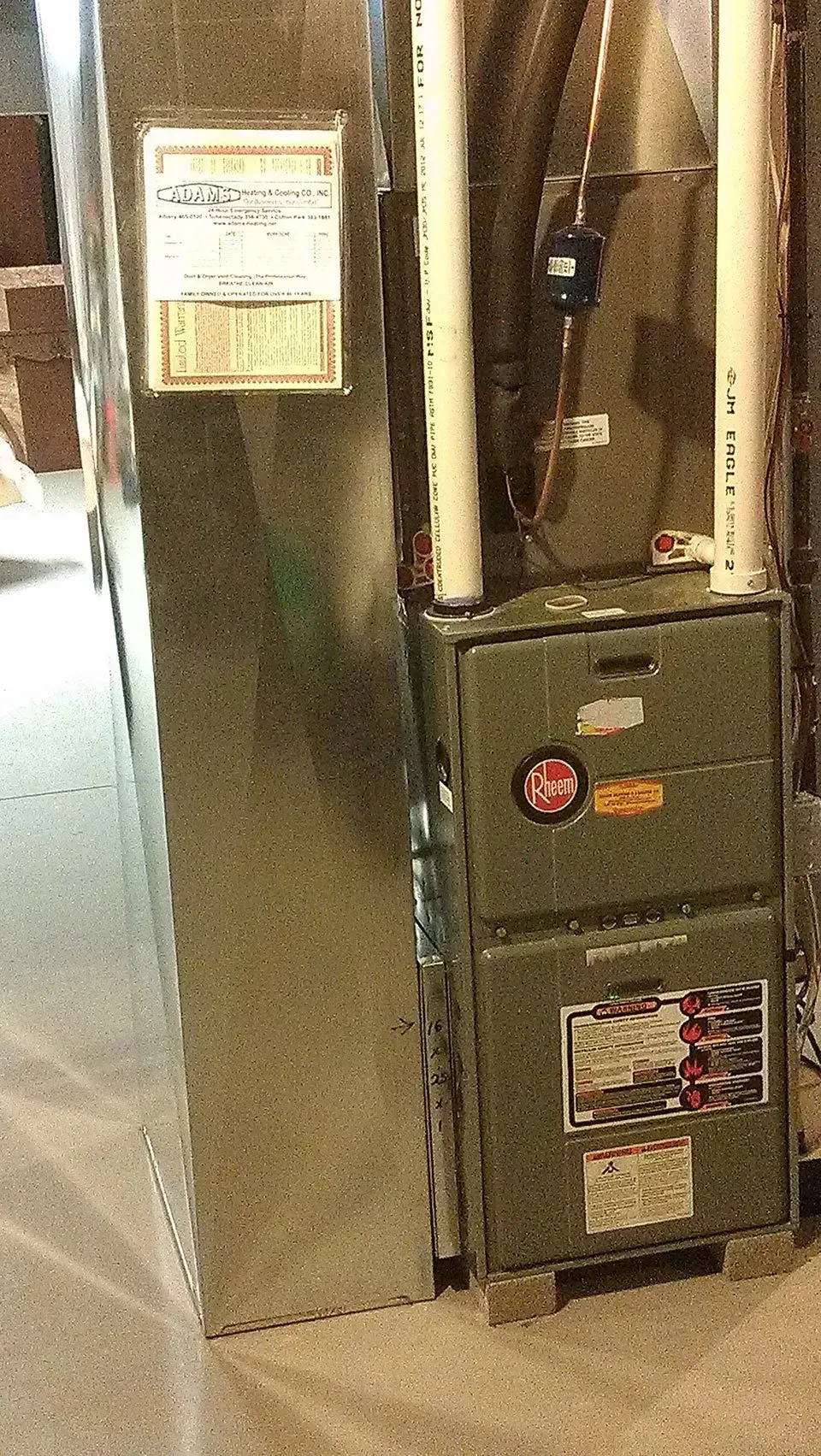 A green furnace unit and a metal air duct standing in a basement, with maintenance labels attached to the front.