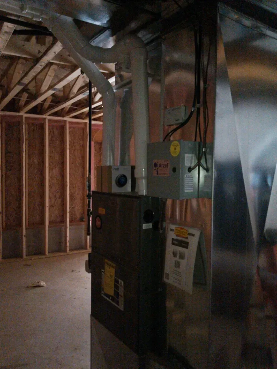A vertical view of a metal HVAC furnace unit installed in an unfinished basement with wood wall studs in the background.