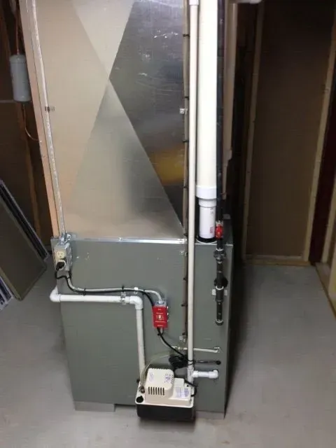 A grey HVAC unit in a utility area with white PVC piping, a condensate pump, and vertical ductwork on a concrete floor.