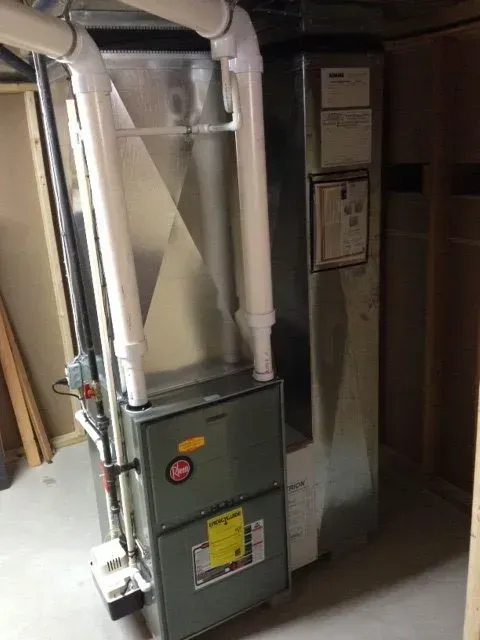 A gray Rheem furnace with white PVC exhaust piping installed in an unfinished basement setting.