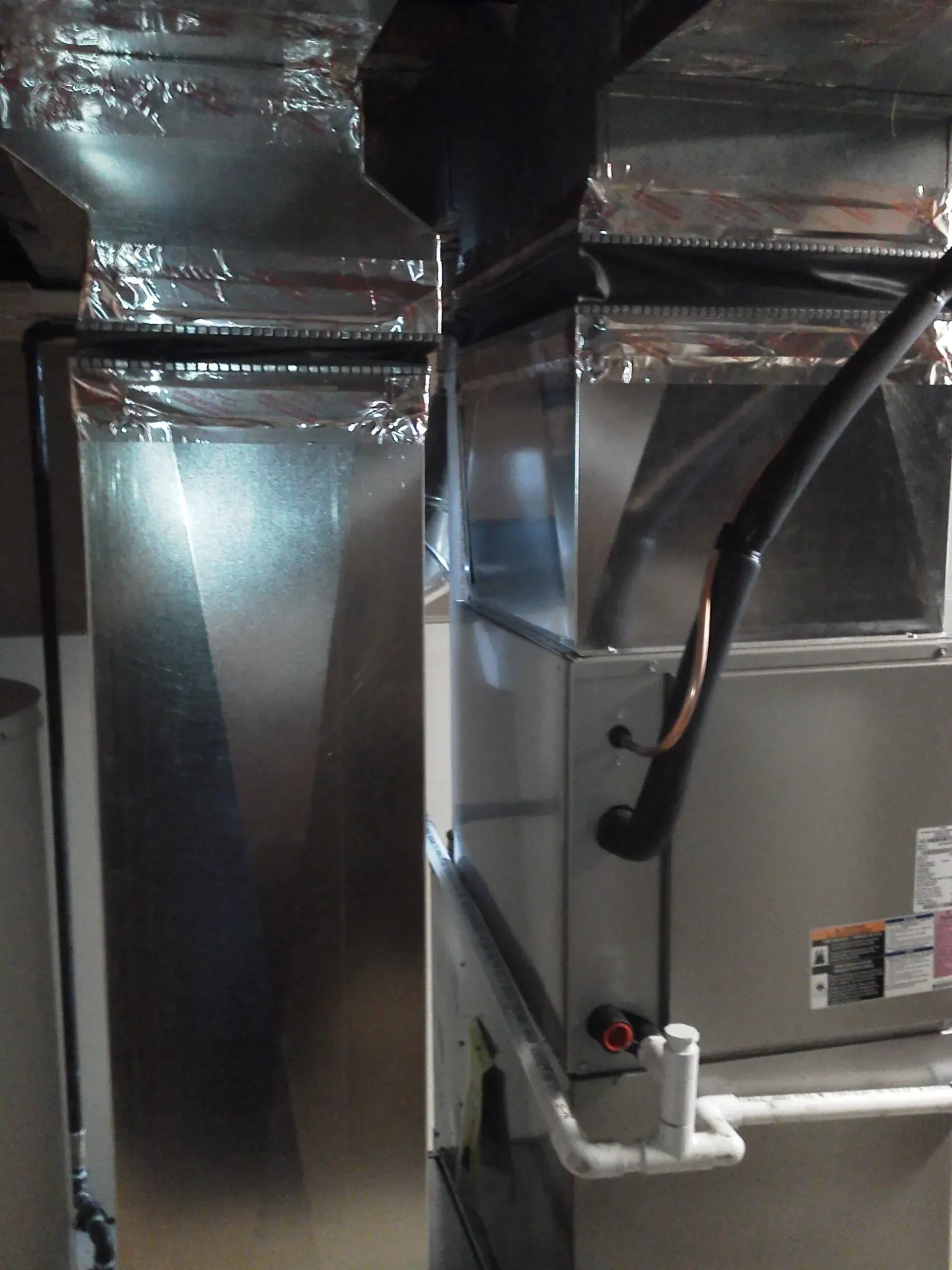 A vertical shot of a silver metal HVAC furnace unit with ductwork connections and attached white PVC piping.