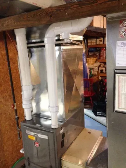 A tall, grey gas furnace with two white PVC pipes extending from the top in a basement or utility room setting.