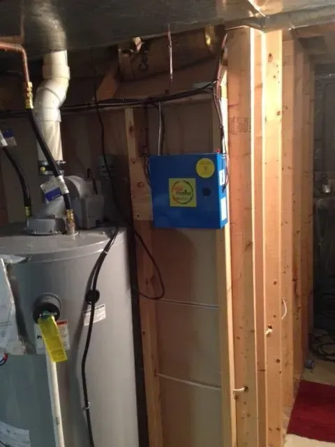 A grey water heater stands next to a blue electronic unit mounted on a wooden basement wall.