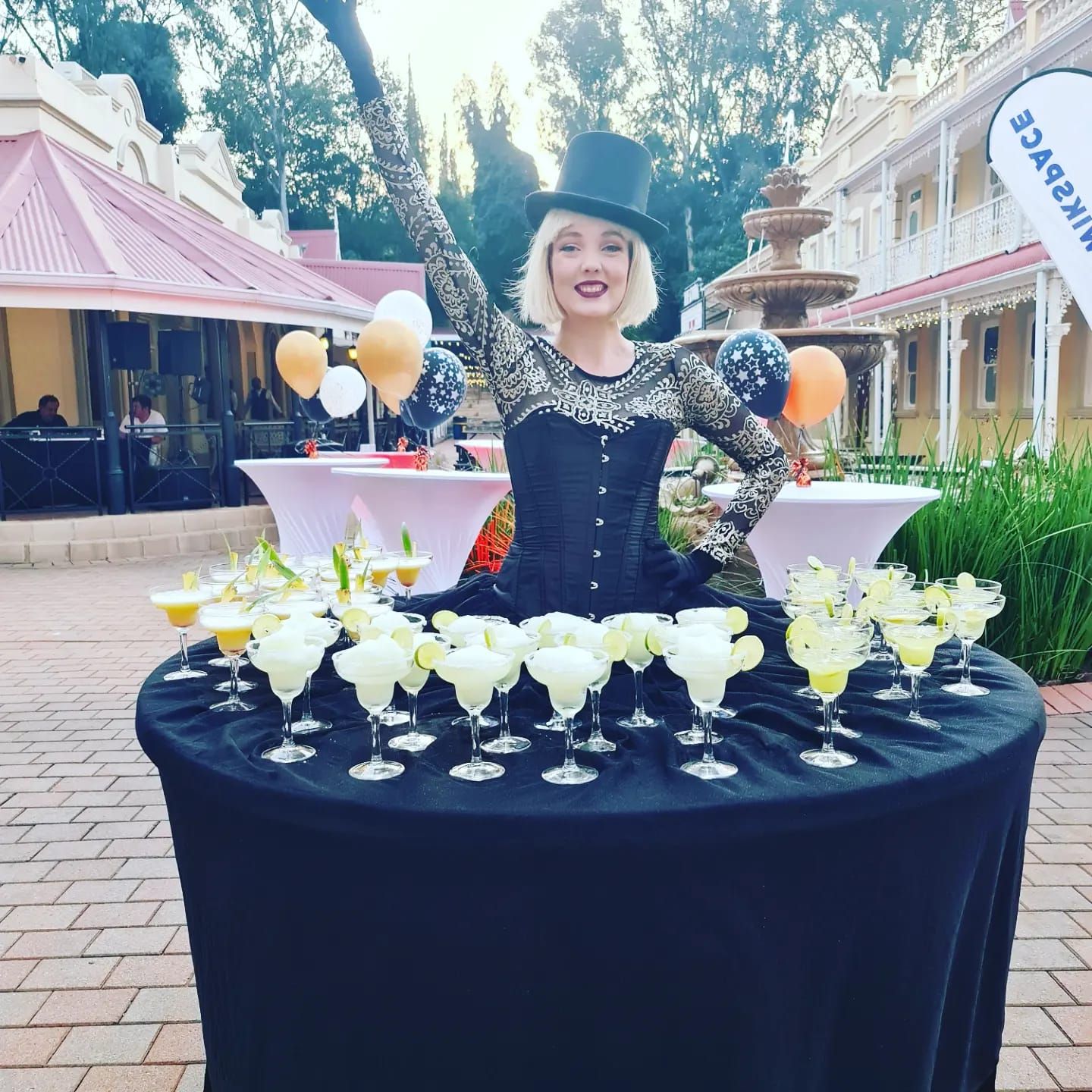 A woman in a top hat stands behind a table filled with glasses of drinks.