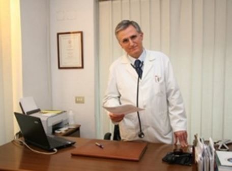 Un medico in camice bianco tiene in mano dei documenti, in piedi dietro una scrivania in uno studio medico.