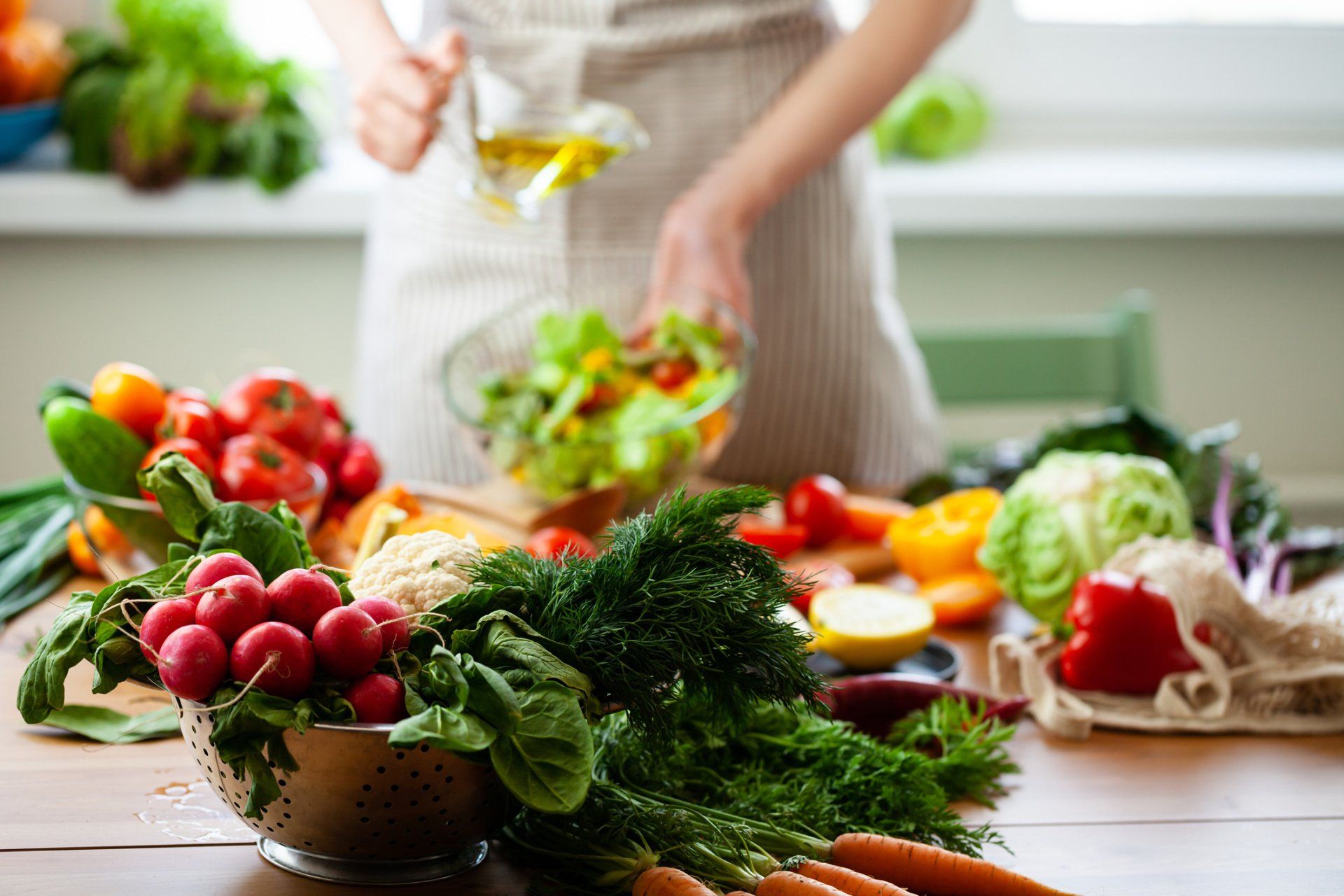 Donna con grembiule che versa olio su un'insalata con verdure fresche e ingredienti su un tavolo di legno.