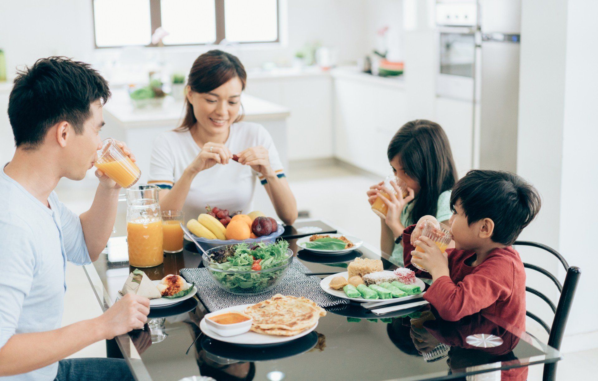 Una famiglia mangia insieme a un tavolo in una cucina luminosa. Bevono succo di frutta e mangiano un'insalata e una macedonia.