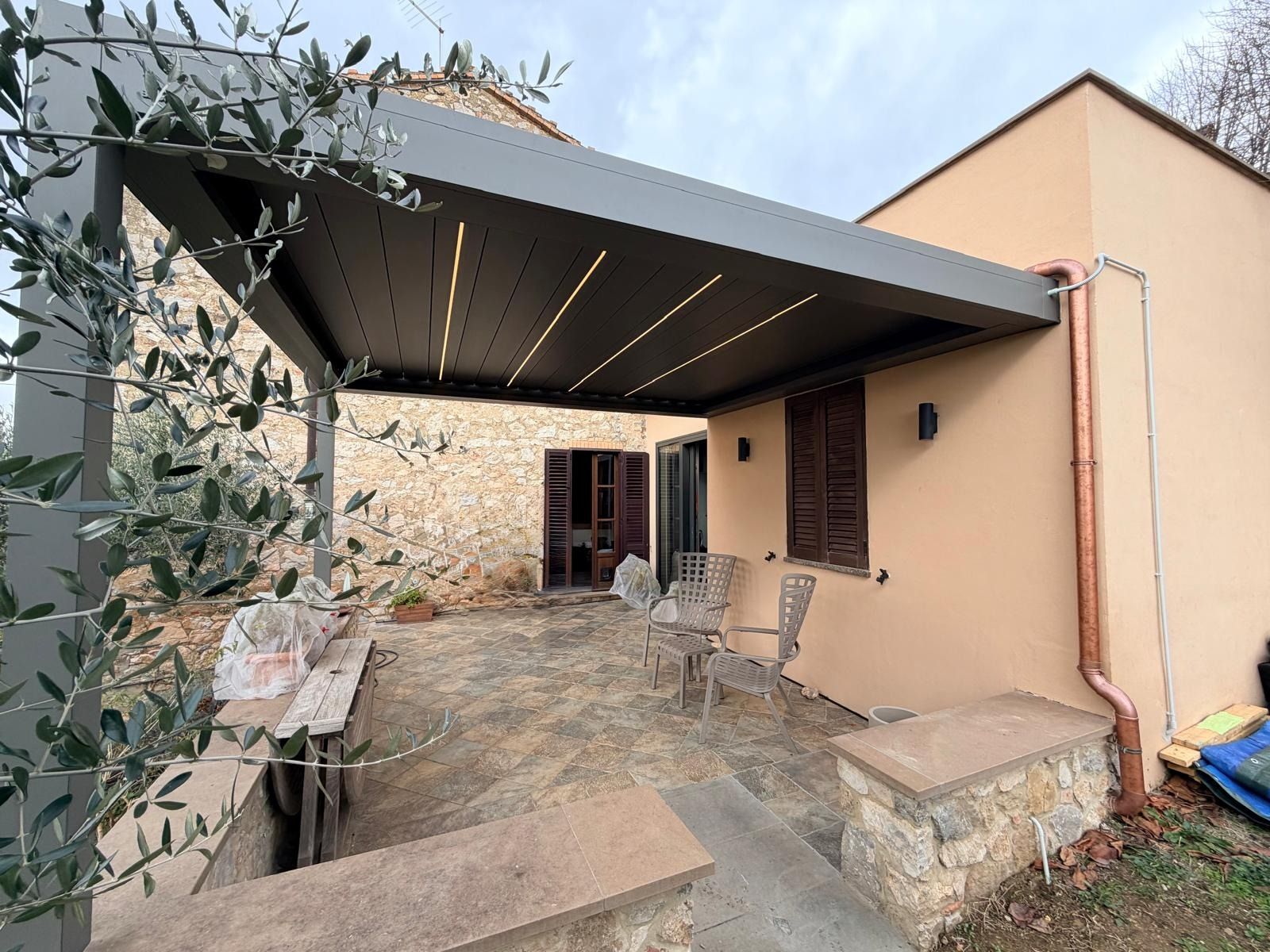 Pergolato che copre un patio accanto a un edificio color marrone chiaro con persiane marroni e grondaia color rame.