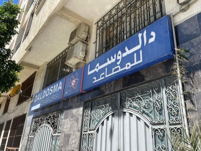 Inscription arabe bleue sur la façade beige d'un bâtiment, au-dessus d'un portail métallique orné.