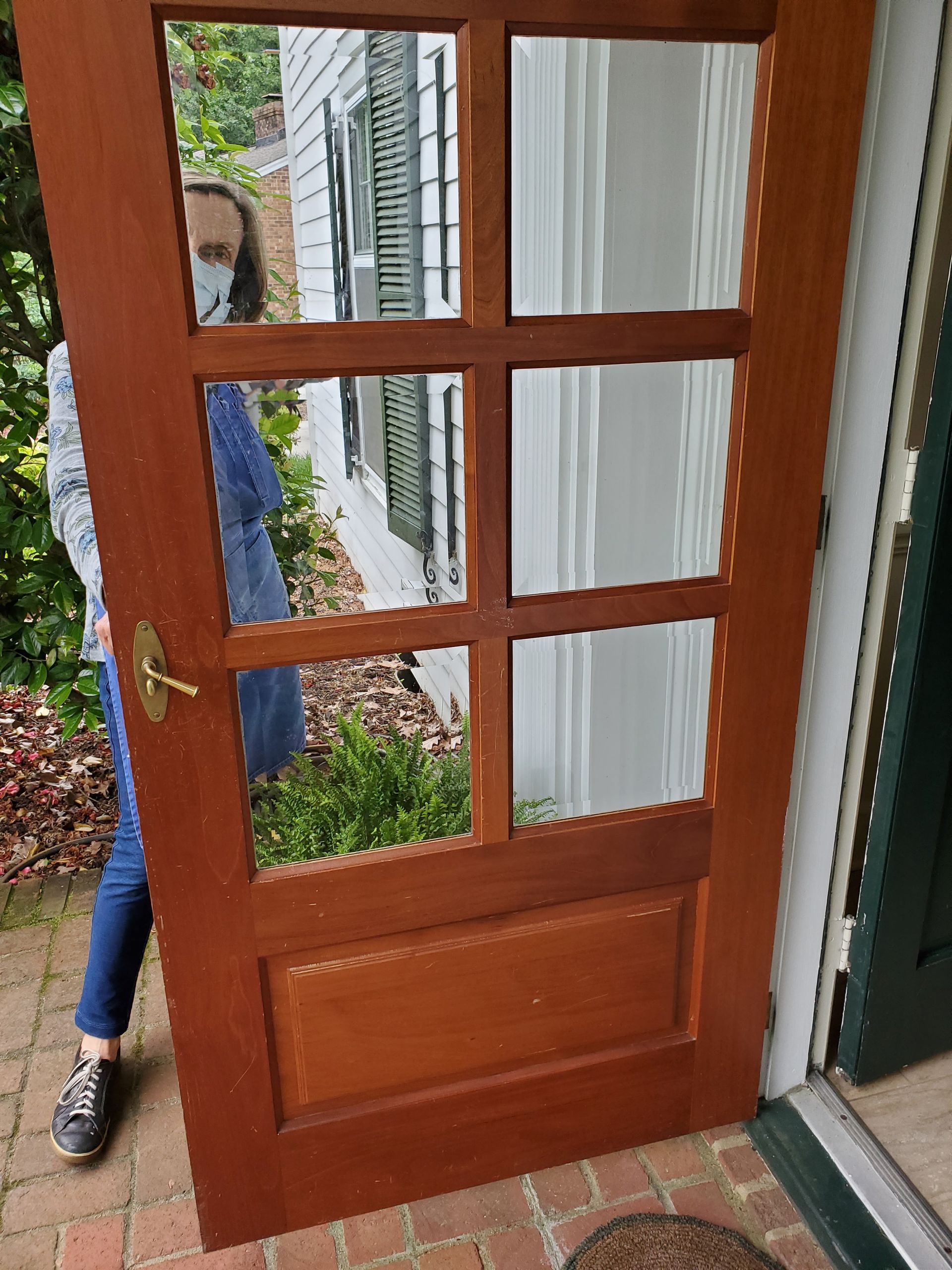 Person wearing a mask stands behind a brown door with glass panels. Brick patio and a white house are visible.