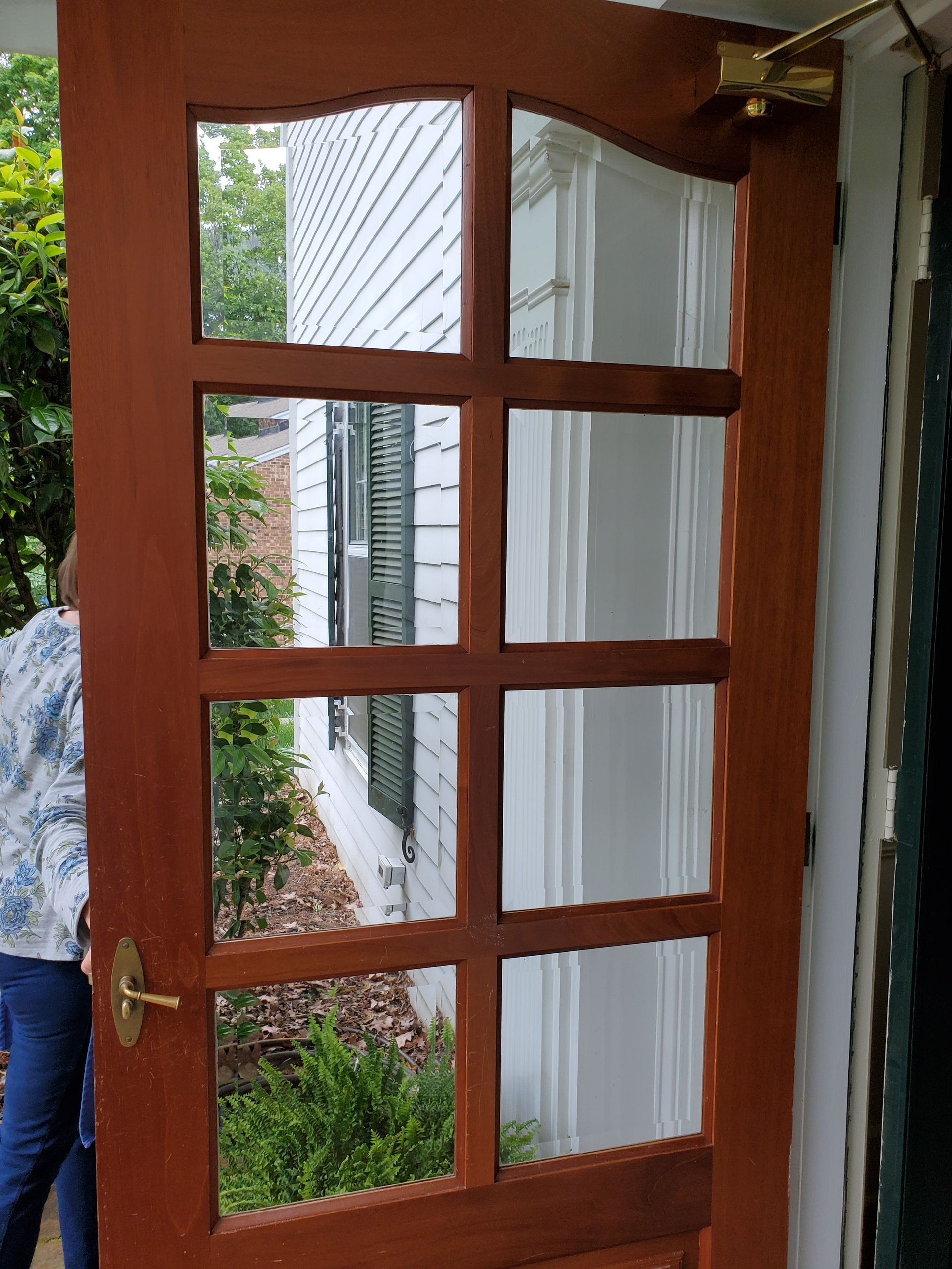 Brown wooden door with glass panes, open to reveal a white house exterior and greenery.