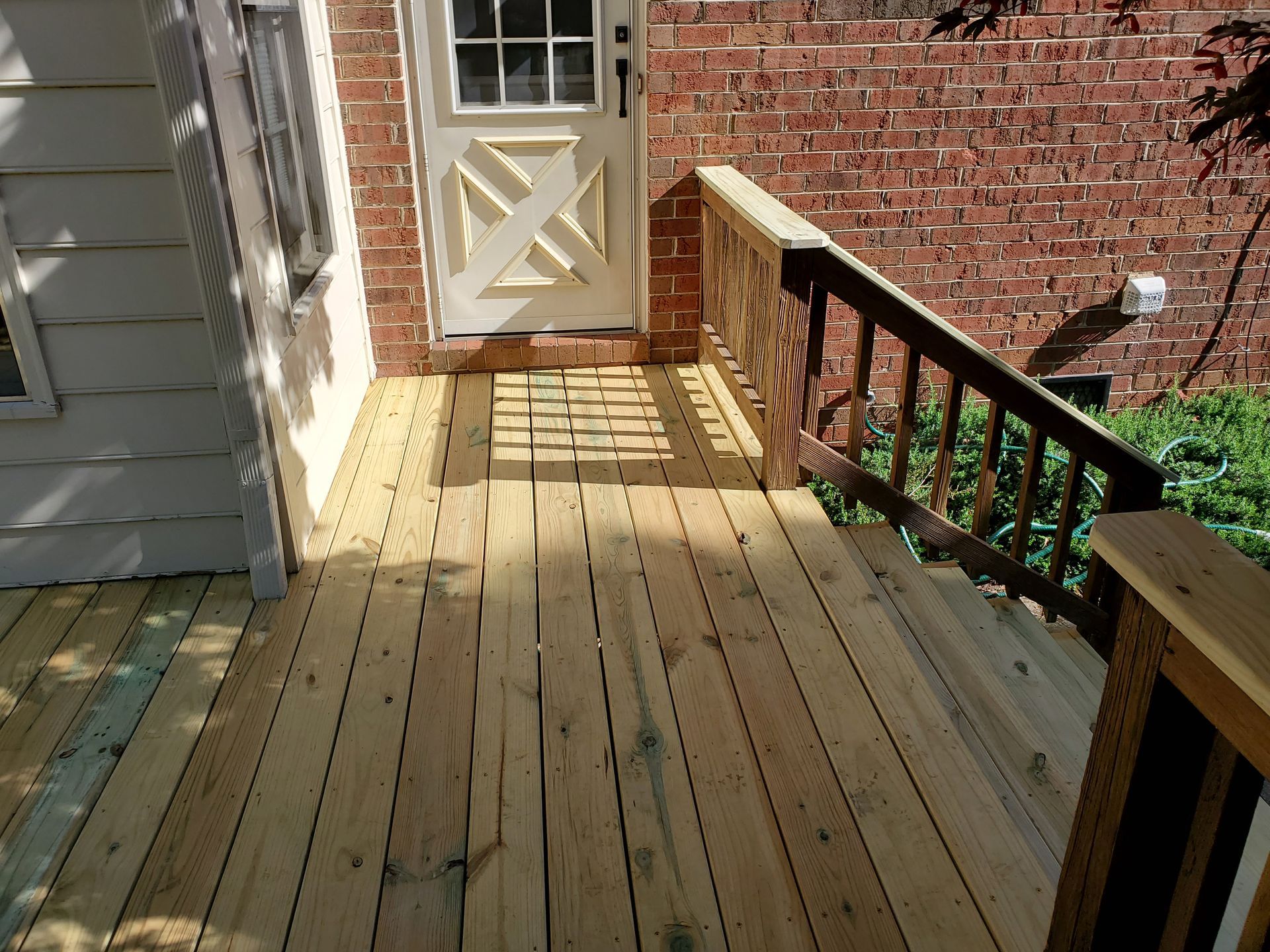 Wooden deck with stairs leading to a door; brick wall and white siding. Handrails.
