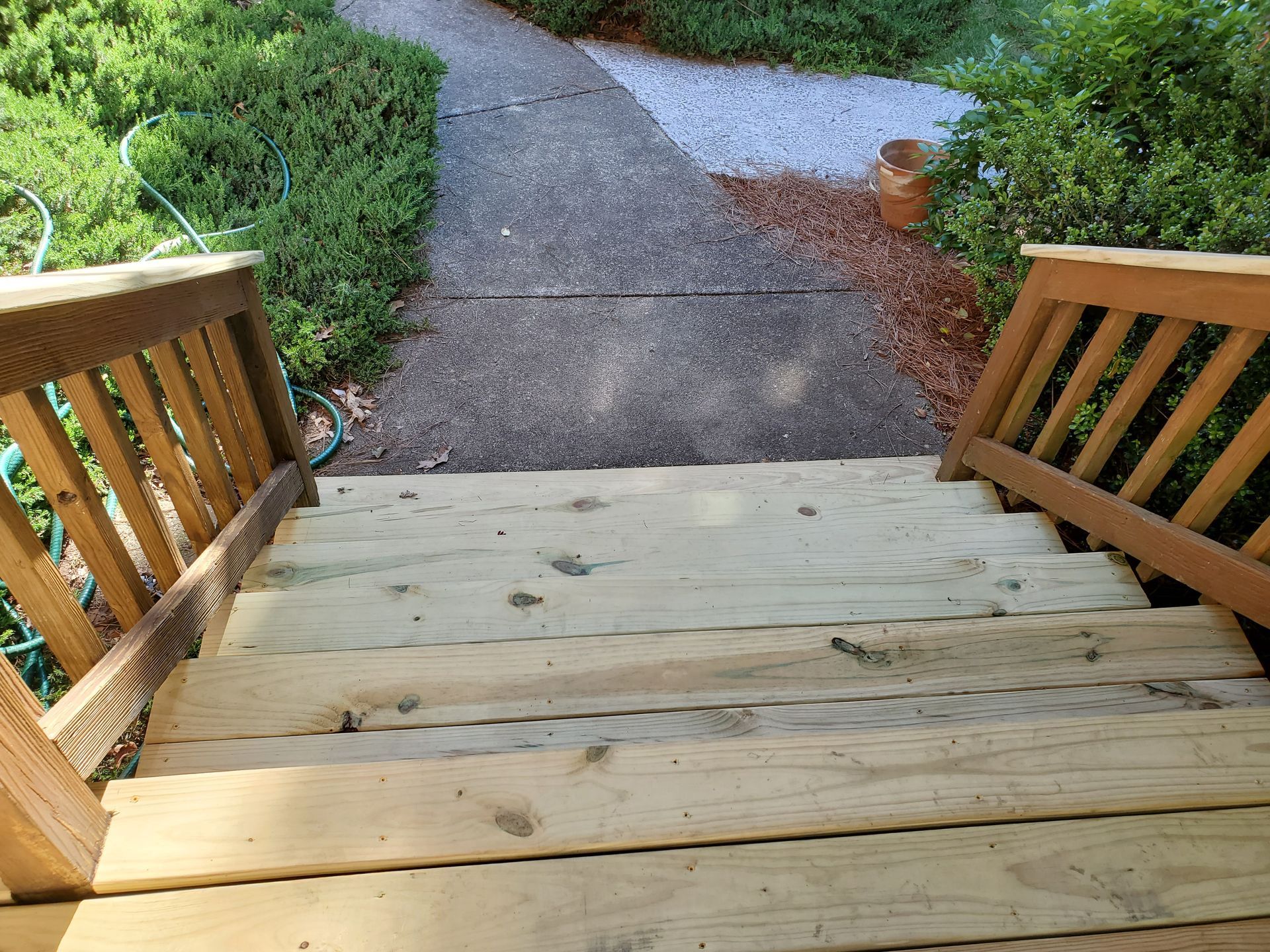Wooden steps leading down to a gray walkway, flanked by wooden railings, and greenery.