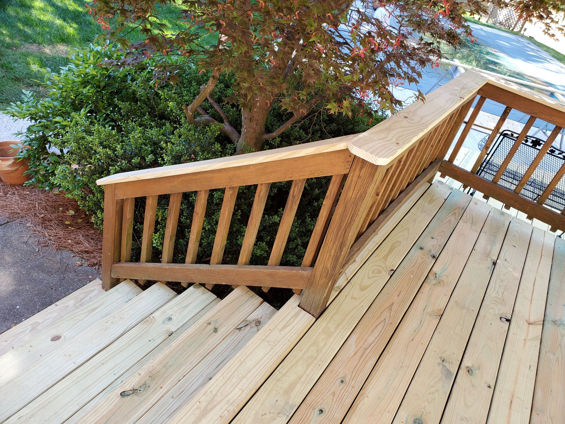 Wooden deck stairs with a brown railing, leading up to a deck near green bushes and a tree.