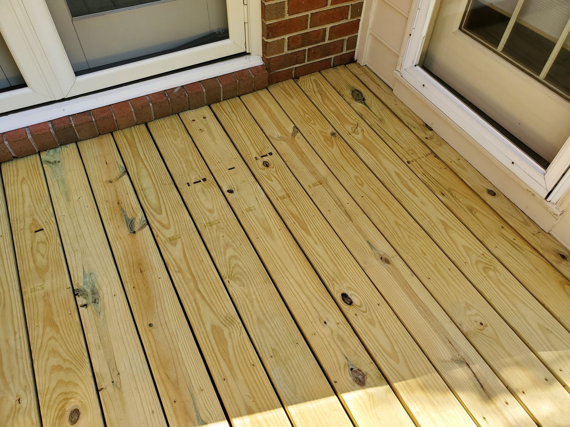 Wooden deck next to brick wall and windows. The deck planks are light brown and run parallel.