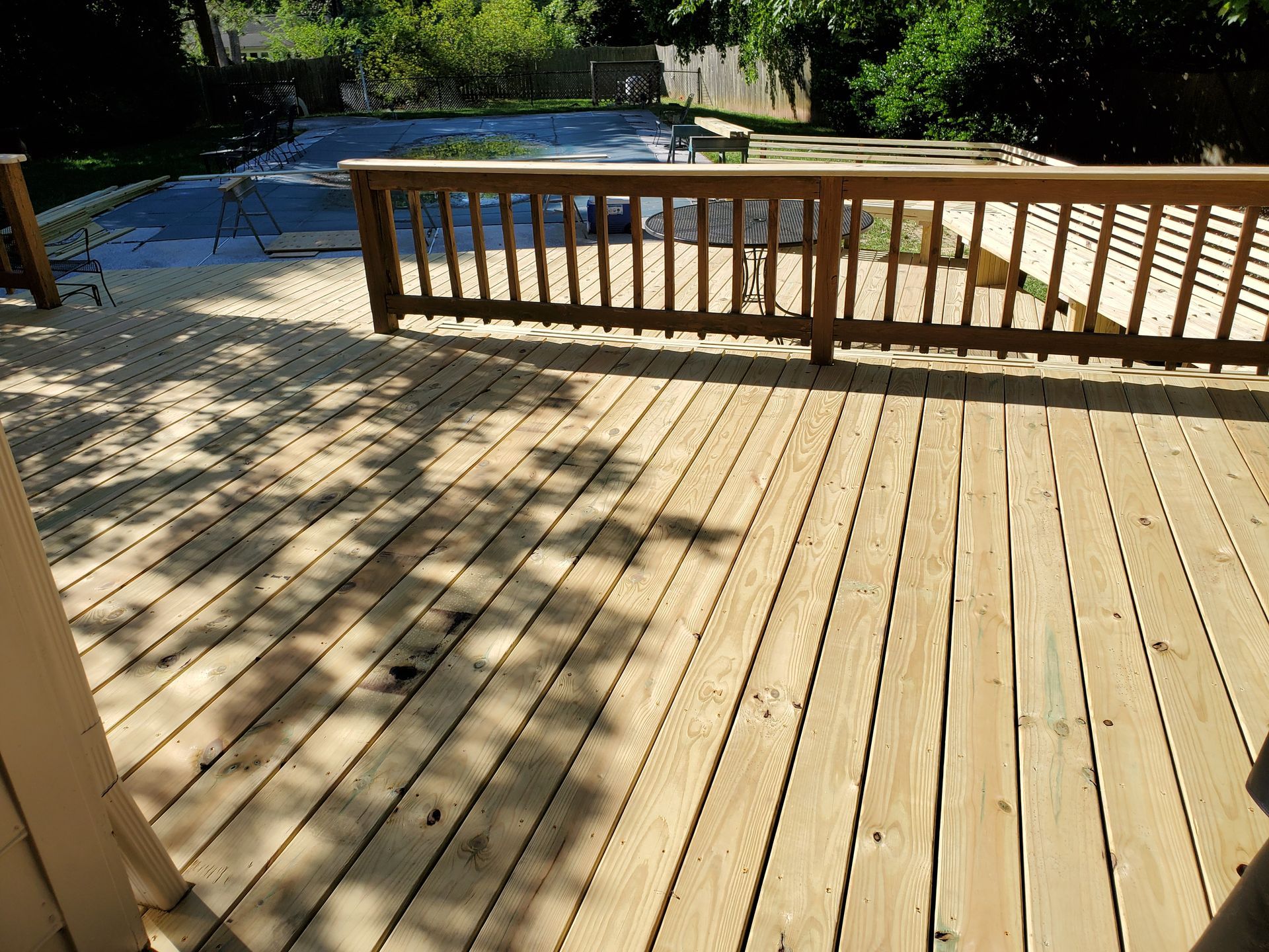 Wooden deck with a railing overlooking a swimming pool, shaded by trees.