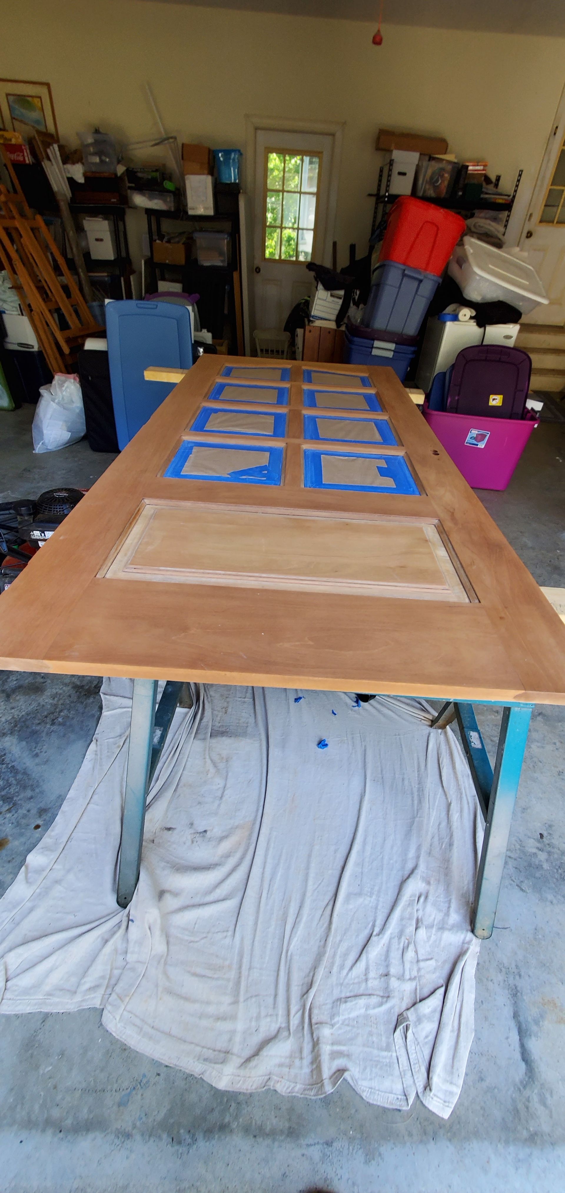 A wooden door is on a work table in a garage. Parts of the door are taped off with blue tape.