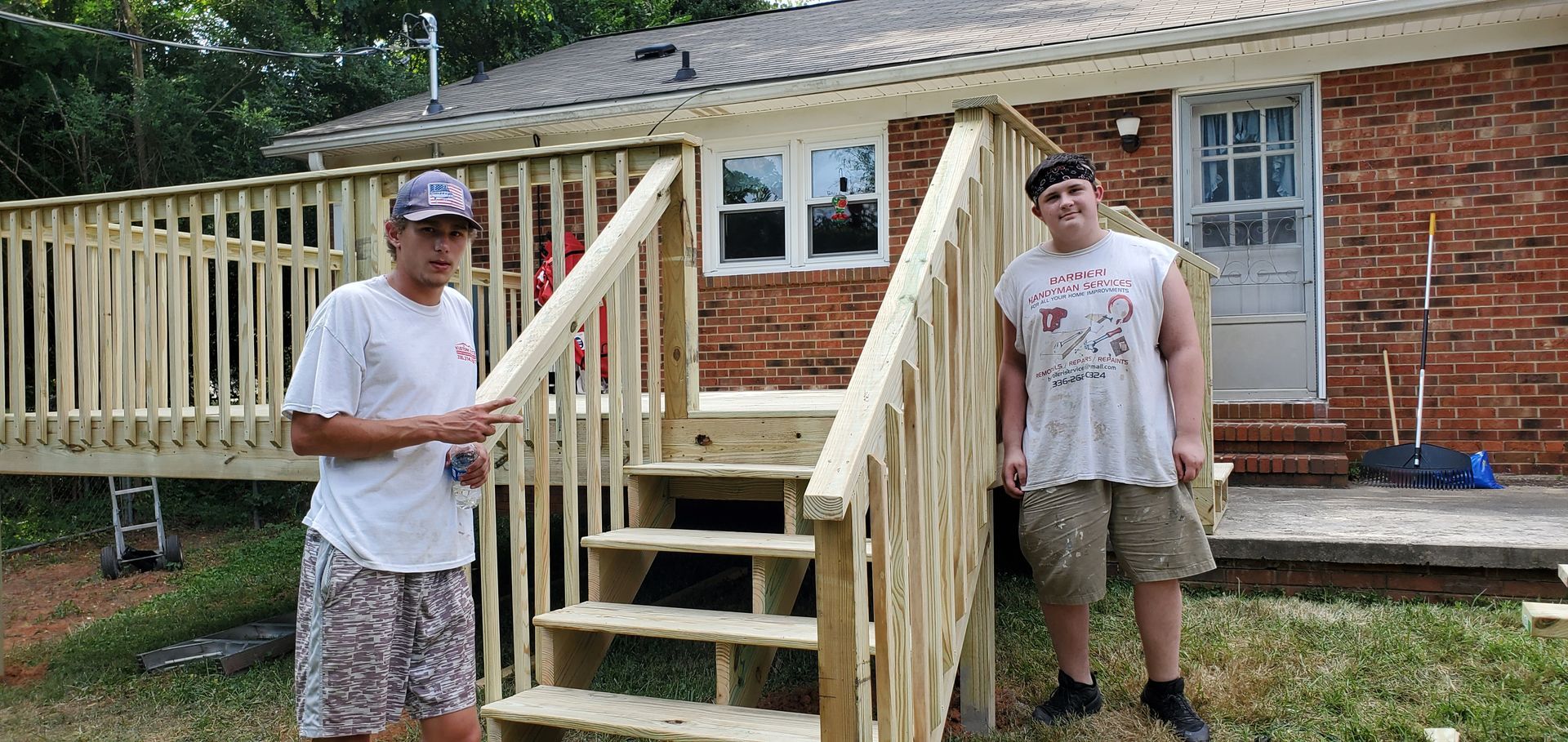 Two people stand on a newly constructed wooden deck, with a brick house as the backdrop.