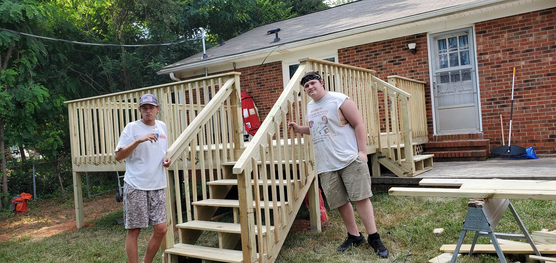 Two people stand near a newly built wooden deck with stairs in front of a brick house.