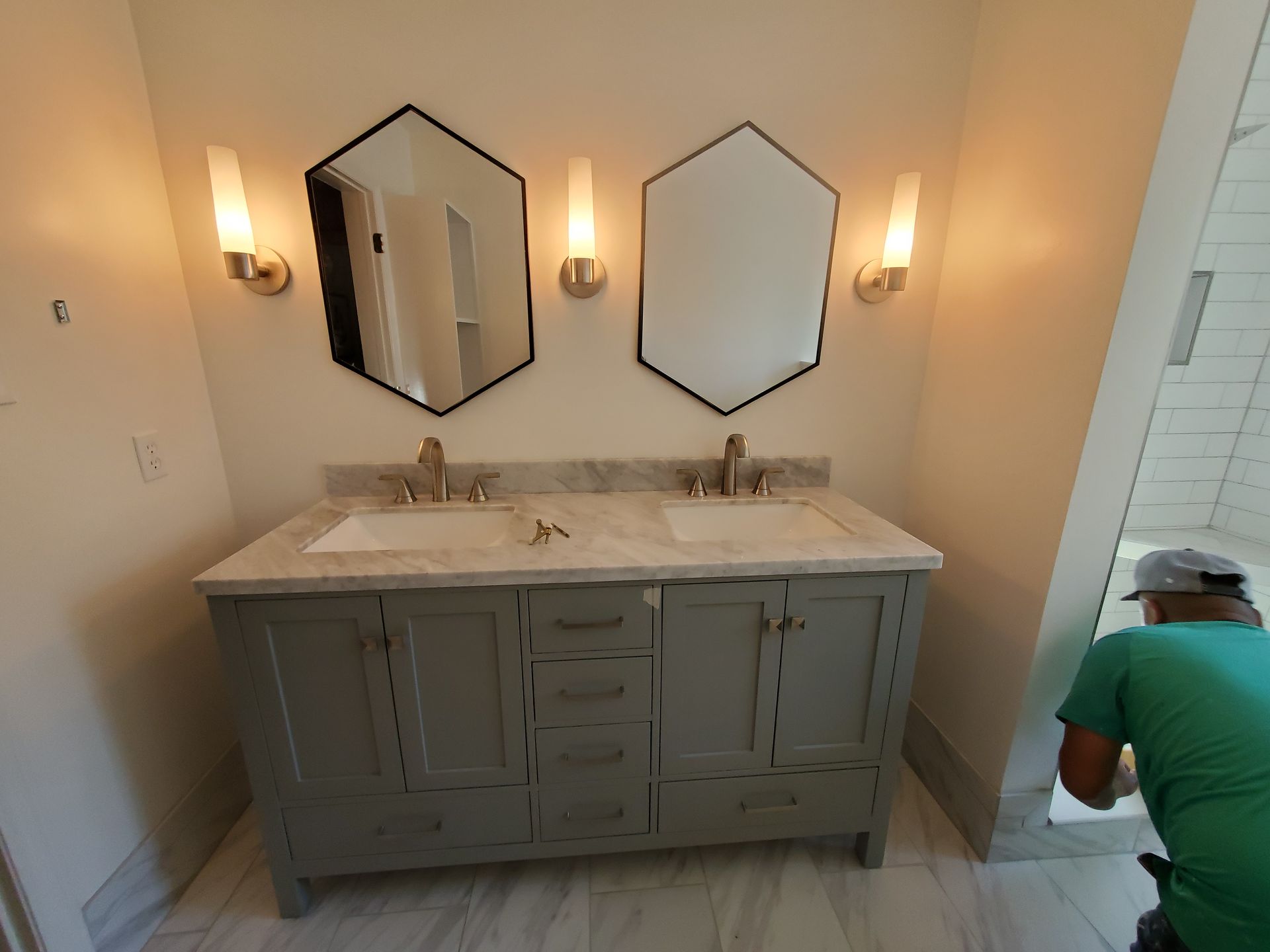 Bathroom vanity with two sinks, hexagon mirrors, sconces, and a person installing tile.