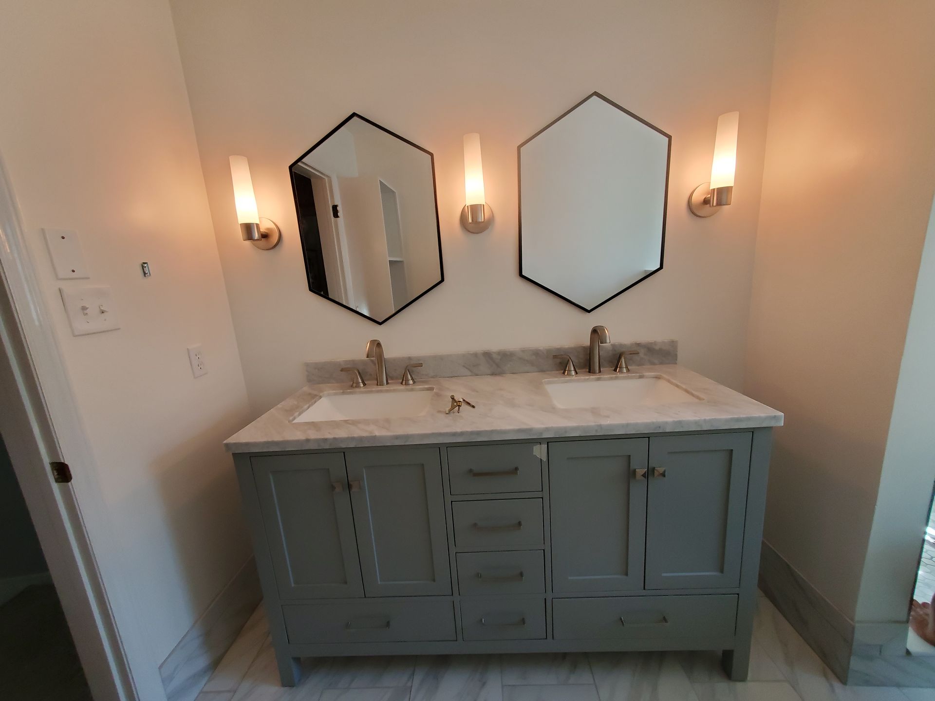 Bathroom vanity with two hexagonal mirrors, gray cabinets, marble countertop, and sconce lighting.
