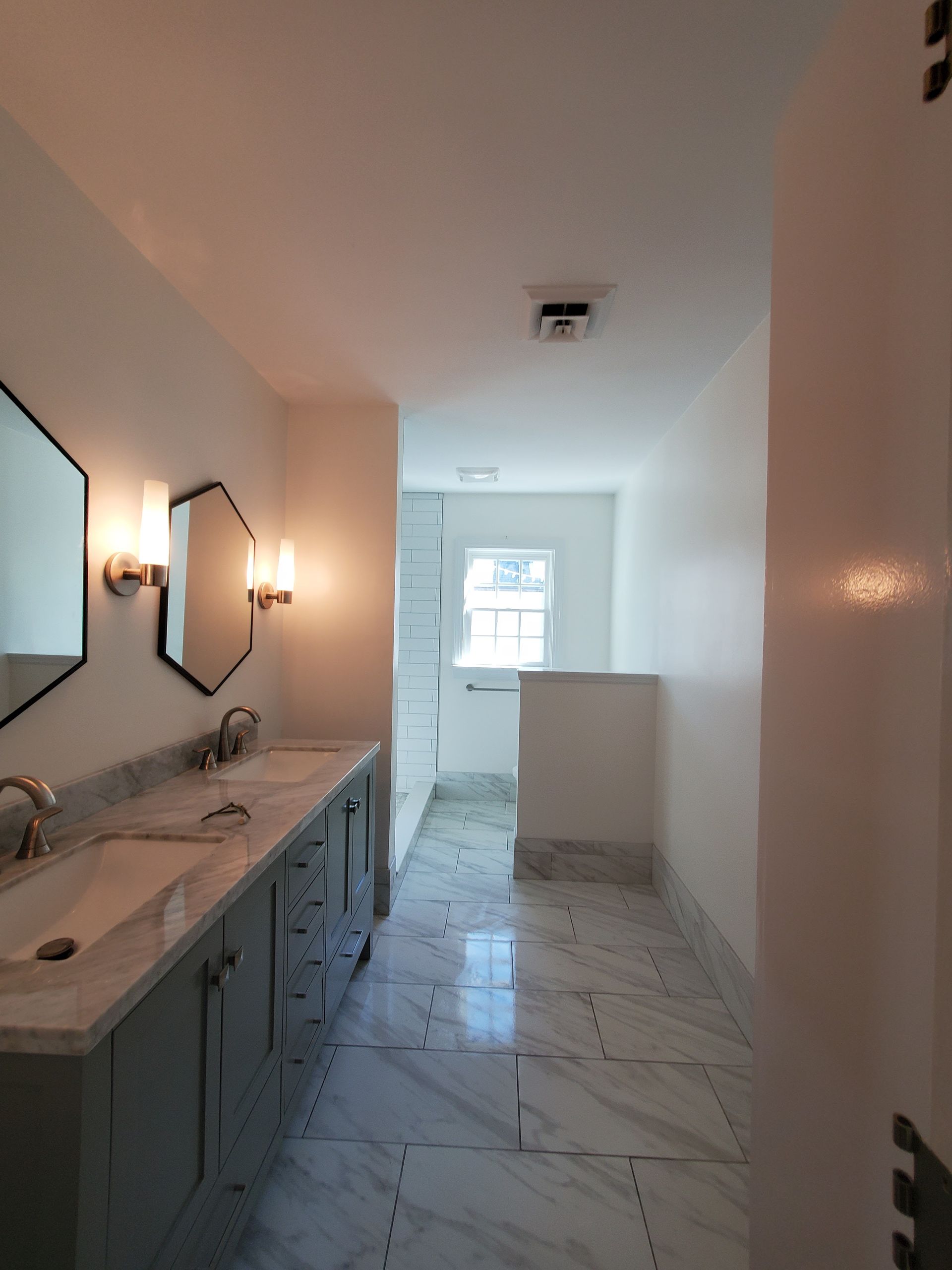 Bathroom with double sinks, gray cabinets, marble floors, and mirrors with sconces.