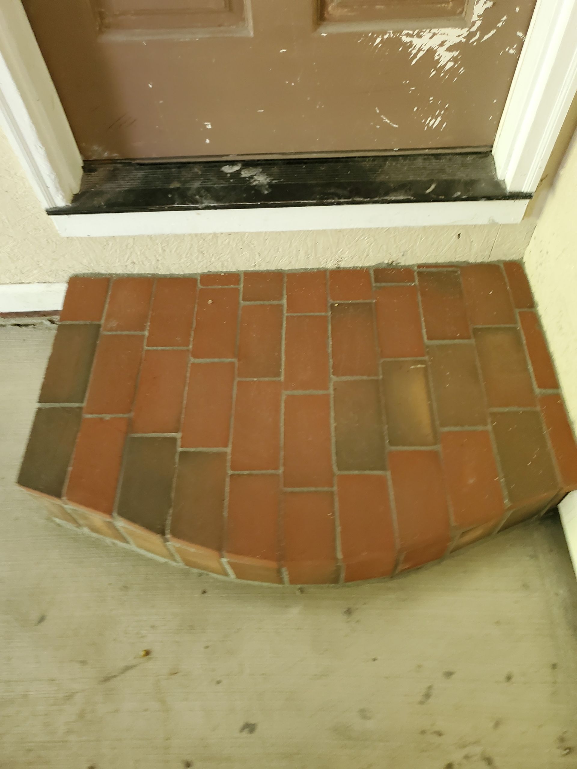 Brick stoop in front of a brown door with peeling paint.  The stoop has a curved front edge and red-brown tiles.