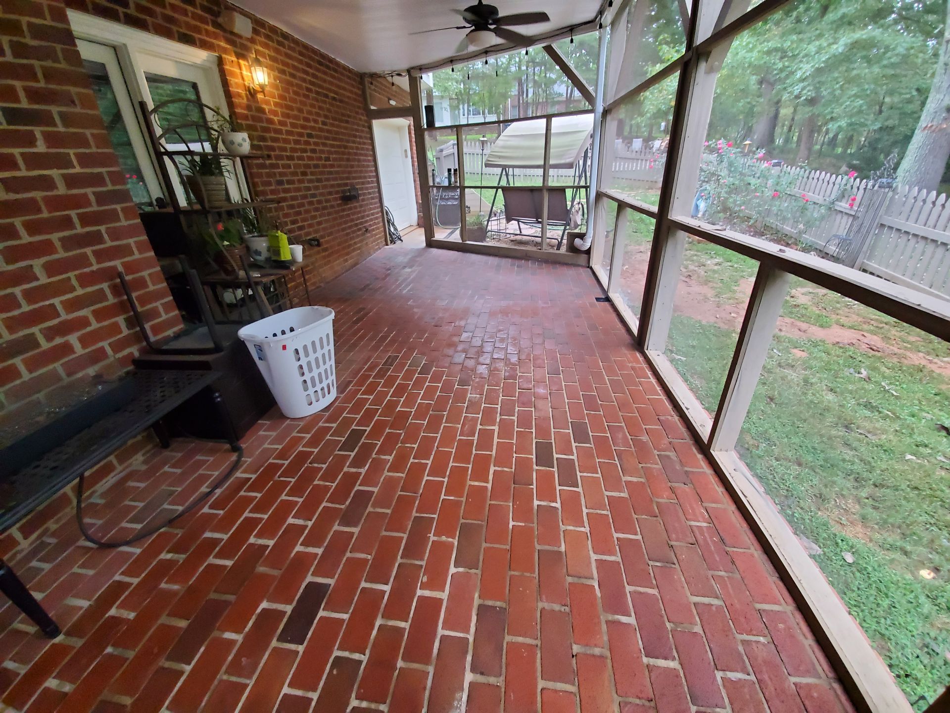 Brick-paved screened porch with view of a yard. A white laundry basket and bench are visible.