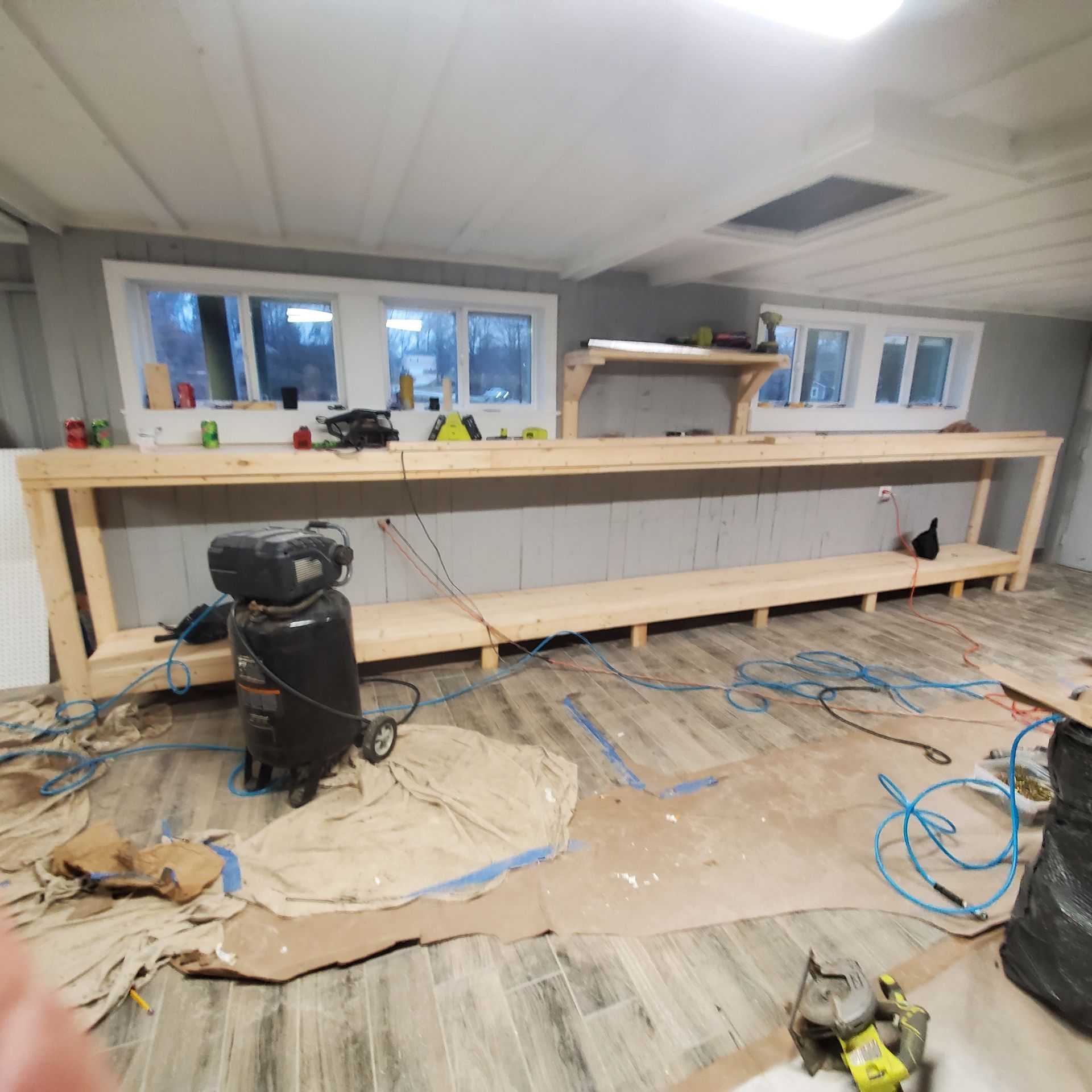 A long, unfinished wooden workbench in a room with windows, an air compressor, and tools; construction in progress.