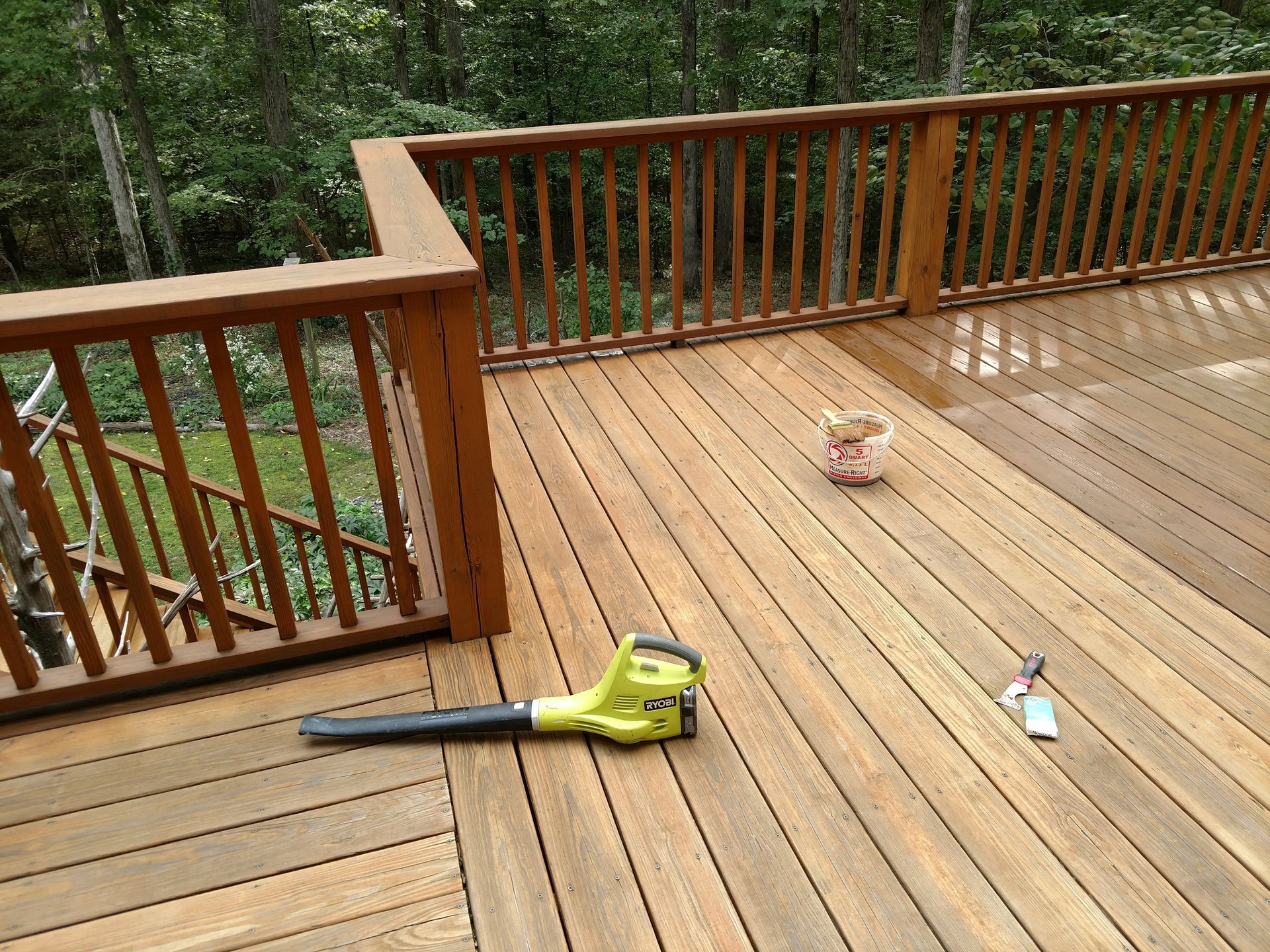 Wooden deck being cleaned with a green leaf blower, with brown railings and a bucket, surrounded by trees.