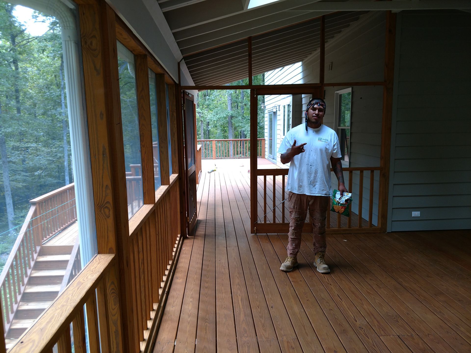 Man standing on a wooden porch, pointing. The porch has a screened roof and overlooks trees.