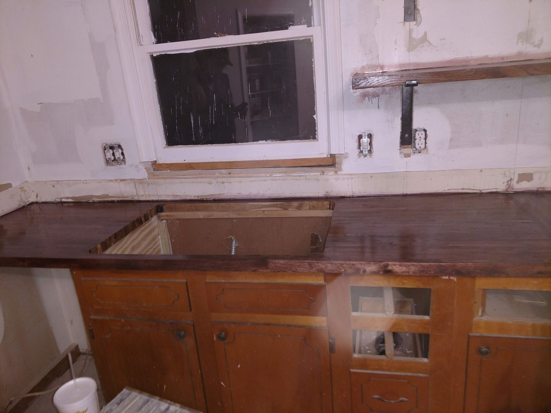Kitchen undergoing renovation with dark countertops, wood cabinets, and a window.