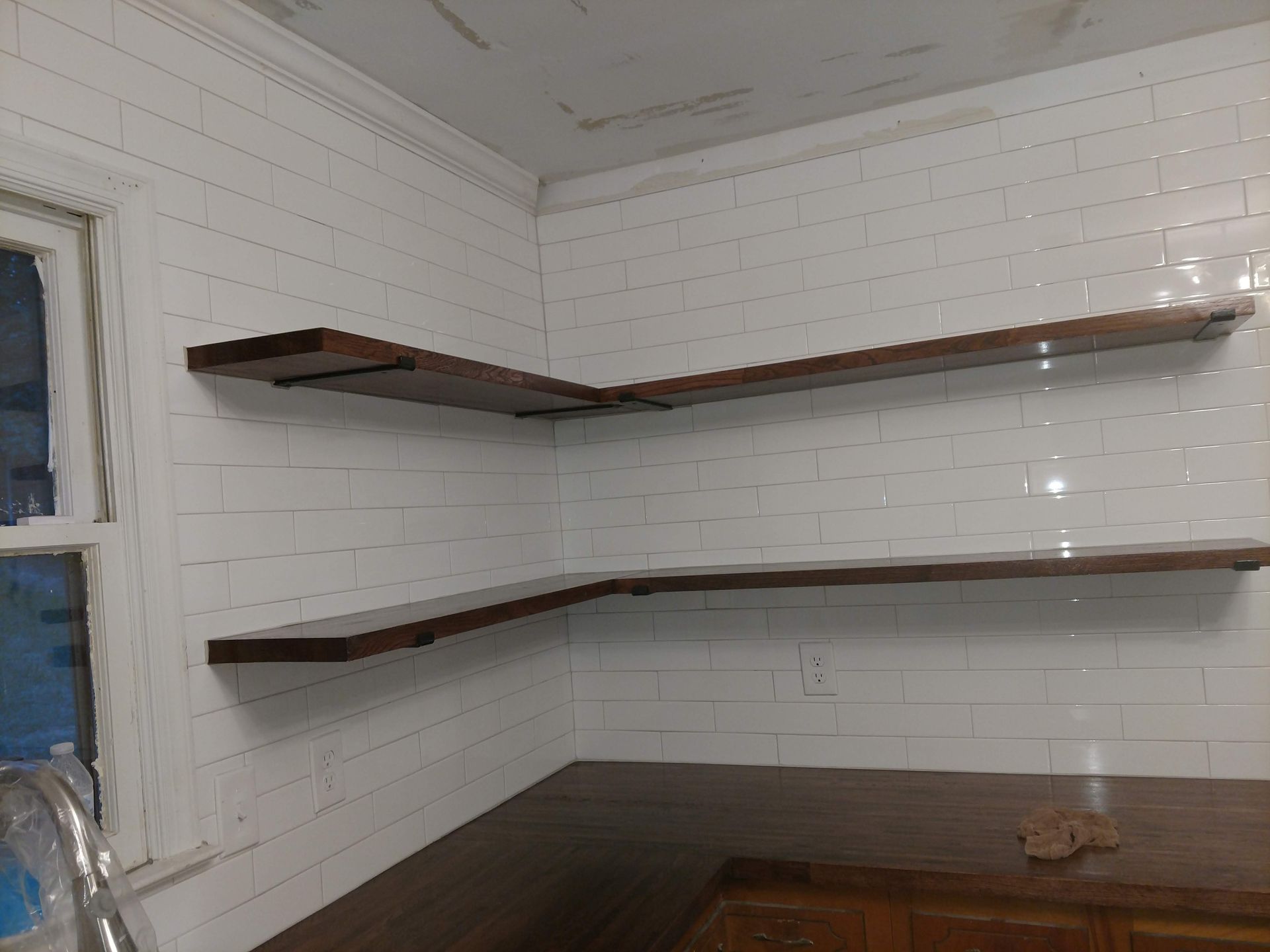 Corner shelves, stained wood, mounted on white tiled wall, above a wooden counter.